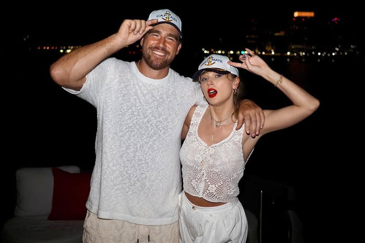 A man and woman wearing matching white caps pose together at night, both adjusting their hats and smiling at the camera.